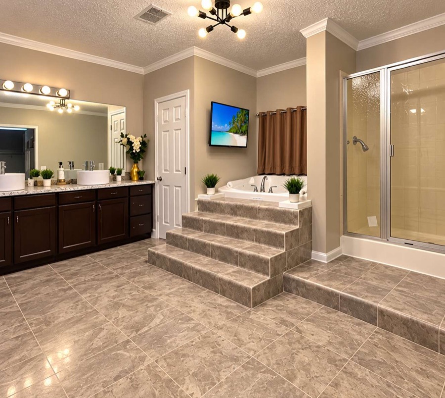The primary bathroom: a jetted tub raised on wide tiled steps, a glass-doored shower alongside, a double vanity with two vessel sinks and a wall-mounted television.