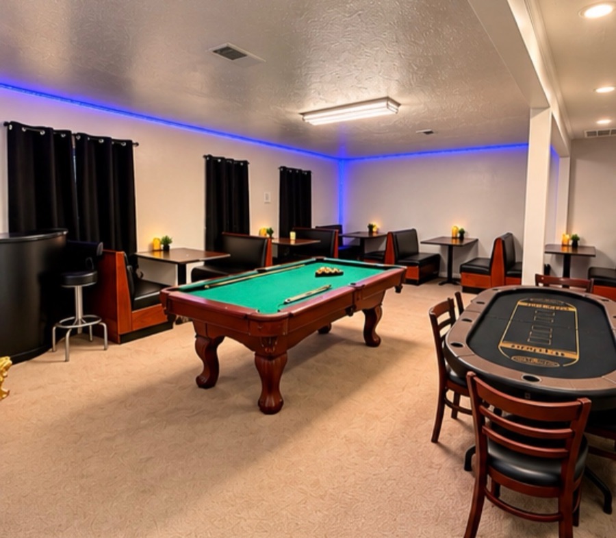 The game room: a pool table with cues laid across the felt, a poker table with chairs, and wooden booth seating under blue cove lighting.