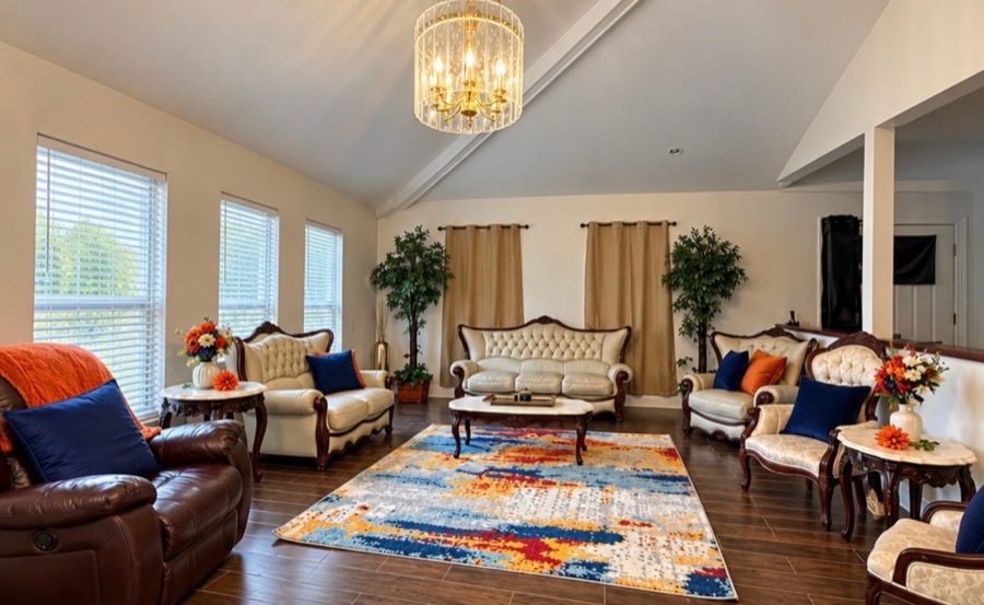 The living room beneath a vaulted ceiling and brass chandelier, with cream tufted sofas and armchairs around a patterned rug and tall curtained windows.
