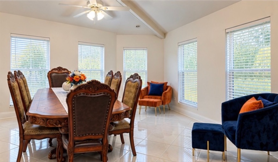 The dining area: a carved wooden table set for eight beneath a vaulted ceiling, with velvet armchairs in orange and blue against a wall of bright windows.