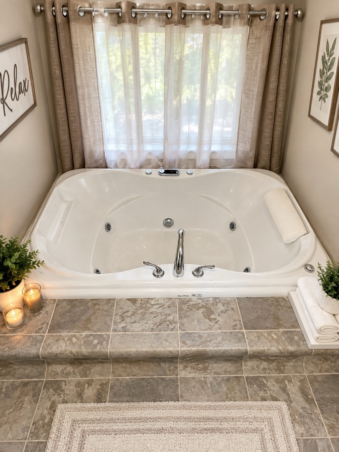 The jetted tub set into a tiled surround beneath a curtained window, with lit candles and rolled white towels on the ledge.